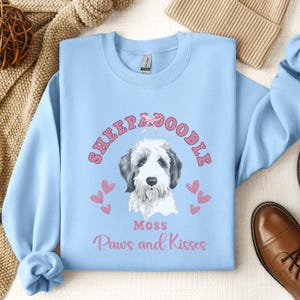 Op de afbeelding: Lichtblauwe trui met een roze en witte afbeelding van een sheepadoodle-hond met de tekst "Sheepadoodle", "Moss", "Paws and Kisses" en roze hartjes.