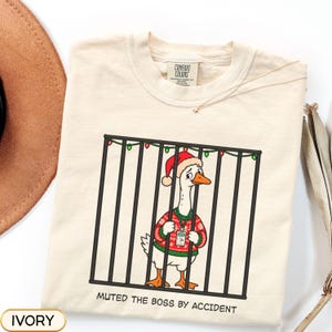 Peut inclure: Un t-shirt ivoire avec une illustration d'une oie portant un bonnet de Père Noël et un pull vert à motifs avec un cordon, debout derrière des barreaux noirs décorés de guirlandes. Le texte en bas indique MUTED THE BOSS BY ACCIDENT.