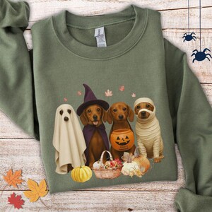 May include: Green sweatshirt featuring a Halloween-themed graphic. The graphic depicts four dachshunds dressed as a ghost, witch, pumpkin carrier, and mummy. A basket of pumpkins and leaves is also shown. The sweatshirt is made of a soft material.