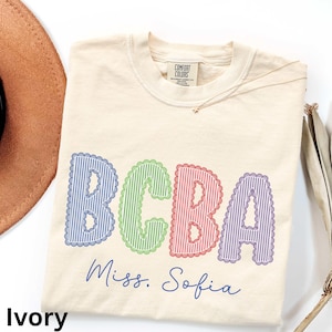 Puede incluir: Camiseta de color marfil con las letras "BCBA" en azul, verde, rojo y morado, con un diseño de borde festoneado. El texto "Miss. Sofia" está debajo de las letras. La camiseta tiene una etiqueta Comfort Colors.