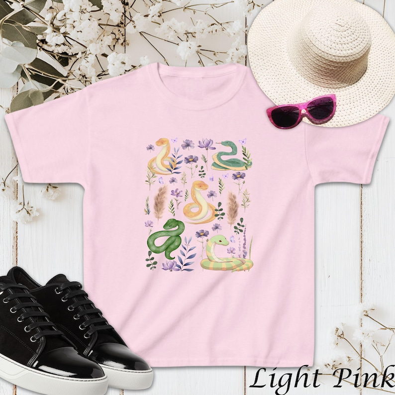 Puede incluir: Una camiseta rosa claro con un dise&ntilde;o colorido de serpientes y flores. Las serpientes son verdes, amarillas y naranjas. Las flores son moradas, rosas y blancas.