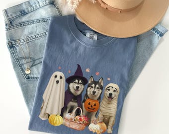 May include: A blue Comfort Colors t-shirt featuring a Halloween-themed graphic of four dogs dressed in costumes. The image includes a ghost, a witch, a dog with a pumpkin bucket, and a mummy. The shirt is paired with light-wash jeans and a tan hat.