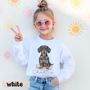 Könnte beinhalten: Weißes Sweatshirt mit einer Illustration eines schwarz-braunen Dackels, umgeben von bunten Blumen. Der Name "Charlie" steht in Schreibschrift darunter. Das Wort "white" befindet sich unten im Bild.
