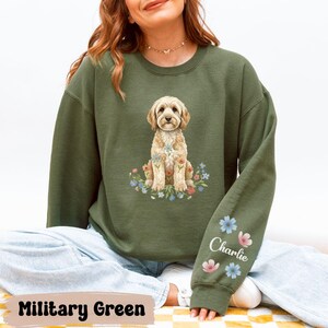 Puede incluir: Sudadera verde militar con una ilustración de acuarela de un perro golden doodle y flores. El perro es el punto central, con el nombre "Charlie" y detalles florales en la manga. La sudadera es de estilo cómodo e informal.