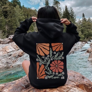 May include: Black hooded sweatshirt with a floral design on the back. The design features orange, red and green flowers with white accents.