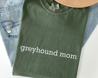 May include: Olive green t-shirt with the text "greyhound mom" in white. The shirt is folded and placed on top of a pair of blue jeans. A tan hat is visible in the upper right corner.