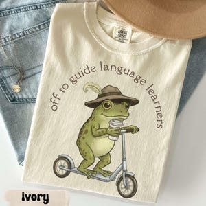 May include: Ivory-colored t-shirt with a frog illustration wearing a hat and riding a scooter while holding a coffee cup. The text "off to guide language learners" is arched around the frog. The word "ivory" is printed at the bottom.
