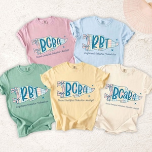 Könnte beinhalten: Fünf kurzärmelige T-Shirts mit Rundhalsausschnitt in Pastellrosa, Hellblau, Salbeigrün, Blassgelb und Creme. Jedes Shirt zeigt eine Grafik eines karierten Wimpels mit dem Text BCBA oder RBT und dem darunter gedruckten vollständigen Jobtitel.