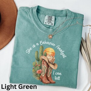 Könnte beinhalten: Ein hellgrünes Kurzarm-T-Shirt mit einer Grafik aus Kaktus, Cowboyhut und Cowboystiefeln. Der Text lautet "She is a Behavior Analyst I can tell". Das T-Shirt hat ein Comfort Colors-Etikett am Halsausschnitt und eine dünne goldfarbene Halskette.