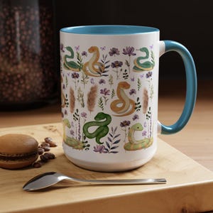 Könnte beinhalten: Weiße Keramiktasse mit blauem Rand und Henkel. Die Tasse zeigt ein sich wiederholendes Muster aus grünen und gelben Schlangen mit floralen und Blattmotiven.