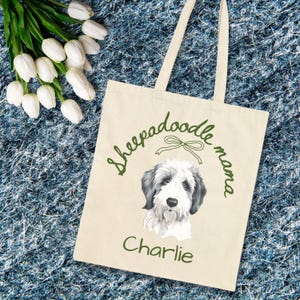 May include: A beige tote bag with the words "Sheepadoodle mama" and the name "Charlie" in green script. The bag features a watercolor illustration of a Sheepadoodle dog with black and white fur.