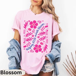 Op de afbeelding: Lichtroze T-shirt met een roze bloemenontwerp en de tekst "Helping little minds bloom." Het woord "Blossom" staat onderaan het shirt. Het shirt heeft een comfortabele, casual stijl.