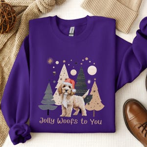 May include: Purple sweatshirt with a Christmas-themed design featuring a dog wearing a Santa hat in front of stylized Christmas trees. The text "Jolly Woofs to You" is printed below the dog.