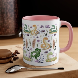 Könnte beinhalten: Eine rosa und weiße Keramiktasse mit einem Muster aus grünen und gelben Schlangen und lila Blumen. Die Tasse hat einen rosa Rand und einen rosa Griff.