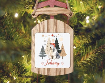 May include: A wooden Christmas ornament shaped like a sled, with a red top and a decorative panel. The panel features a dog wearing a Santa hat, with Christmas trees and the name "Johnny". The ornament hangs from a natural twine string.