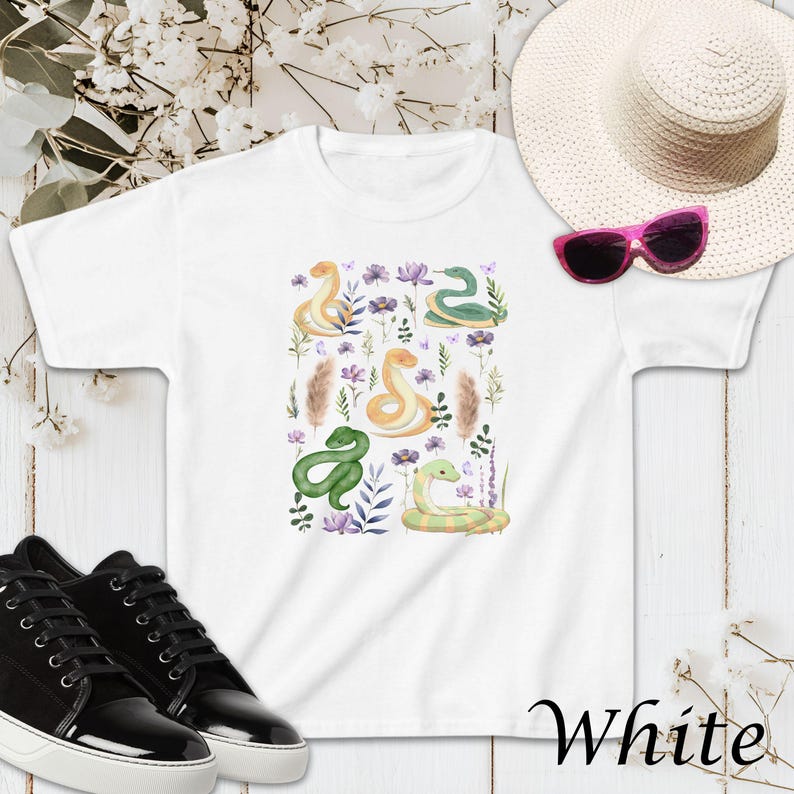 Puede incluir: Camiseta blanca con un estampado colorido de serpientes y flores. El estampado presenta serpientes verdes, amarillas y moradas con flores y hojas en tonos de morado, rosa y verde.
