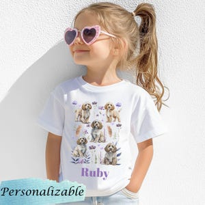 May include: White t-shirt with a personalized name "Ruby" printed in purple. The shirt features a repeated pattern of brown and white dogs with purple flowers and greenery.