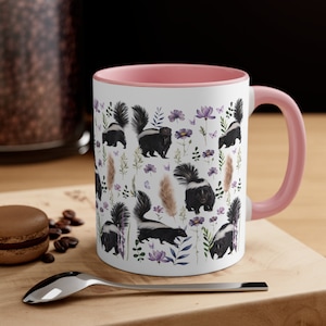 May include: A white ceramic mug with a pink rim and handle. The mug features a repeating pattern of black and white skunks with purple flowers and green leaves on a white background.