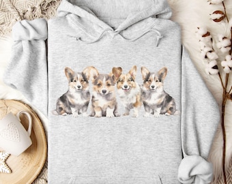 May include: A light gray hoodie featuring a watercolor illustration of four corgi puppies. The puppies are in various shades of brown, gray, and white. The hoodie has a drawstring hood and a kangaroo pocket.