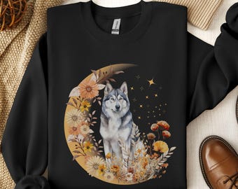 Op de afbeelding: Zwart sweatshirt met een grijze wolf die op een halve maan zit, omringd door gele en oranje bloemen. De maan is goud en heeft een sterrenhemel als achtergrond.