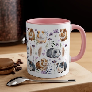 May include: White ceramic mug with a pink rim and handle. The mug features a repeating pattern of watercolor illustrations of guinea pigs and flowers. The guinea pigs are brown, black, and white.