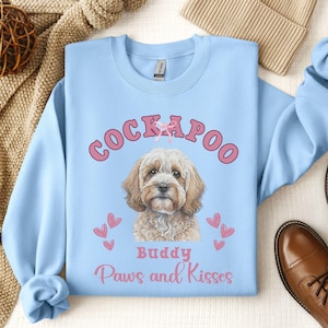 May include: Light blue sweatshirt with a pink Cockapoo dog illustration. The dog is wearing a pink bow and the text "Buddy Paws and Kisses" is printed below the dog.