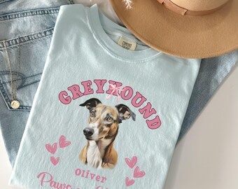 May include: Light blue t-shirt with a pink and white graphic of a Greyhound dog with the text "Greyhound" in pink letters, "Oliver" in black letters, and "Paws and Kisses" in black letters.
