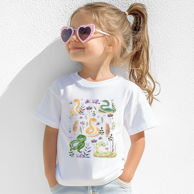 Puede incluir: Camiseta blanca con un estampado colorido de serpientes y flores. Las serpientes son verdes, amarillas y marrones. Las flores son moradas, rosas y blancas.