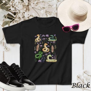 Puede incluir: Camiseta negra con un estampado colorido de serpientes y flores. El estampado presenta serpientes verdes, amarillas y marrones con detalles florales.