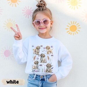 Op de afbeelding: Een jong persoon draagt een witte sweatshirt met een aquarelontwerp van puppy's en bloemen. Het woord "white" staat onderaan de afbeelding. De achtergrond is een muur met geschilderde zon- en bloemontwerpen.