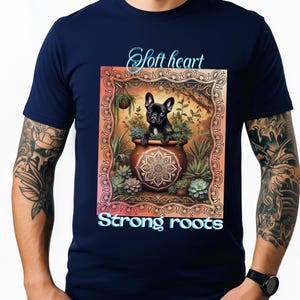 May include: Navy blue t-shirt featuring a graphic of a black French Bulldog in a decorative pot surrounded by plants. The image includes the text "Soft heart" and "Strong roots" in a light blue font.