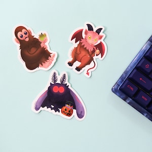 May include: Three colourful stickers featuring a pink devil holding a candy cane, a black moth with red eyes holding a jack-o-lantern, and a brown monkey holding an apple.