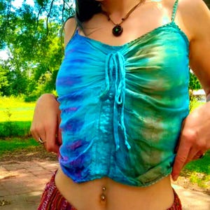 May include: A tie-dye tank top in shades of blue, green, and purple. The top features spaghetti straps, a gathered front, and a drawstring detail. The garment is made of a lightweight fabric, perfect for warm weather.