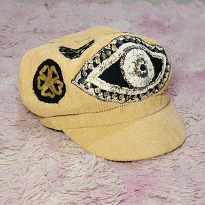 May include: A light tan corduroy newsboy cap featuring a large embroidered eye design in black, white, and gold thread. To the left of the eye is a circular black patch with a gold floral embroidery, and a small black leaf-shaped accent above.
