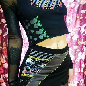 May include: Black crop top with mesh sleeves and a patchwork skirt. The top features an oval patch with colorful embroidery. The skirt has a patchwork design with gray, black, and patterned fabrics. The outfit has a unique, artistic style.