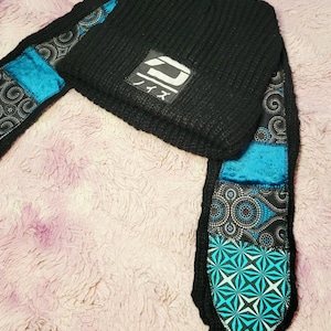 May include: A black ribbed knit beanie with two long scarf-like tails attached. The beanie features a rectangular patch with white Japanese text and a stylized graphic. The tails are decorated with patchwork panels in black, white, and teal blue.