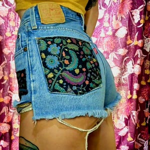 May include: Light blue denim shorts with frayed edges and decorative floral patches on the back pockets. The patches feature a dark background with colorful floral and paisley designs. A Levi's tag is visible.