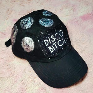 May include: A black baseball cap featuring a rectangular patch with the white text DISCO BITCH. The cap is decorated with several circular, reflective, silver-toned sequined patches arranged across the crown and side panels.