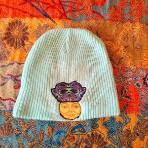 May include: A light blue knitted beanie with a colorful embroidered design of a face with flowers on top.