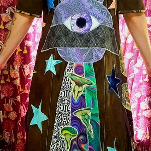 May include: Brown dress with short sleeves, featuring an alien eye design and star accents. The dress has a patchwork design with mushroom and geometric patterns in various colors, creating a unique and artistic look.