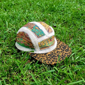 May include: A five-panel cap featuring a patchwork crown with white fabric seams and colorful abstract patterns in shades of green, yellow, and brown. The flat brim is covered in a brown and black leopard print pattern with visible stitching.