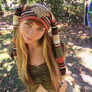 May include: A long, striped knit hat featuring horizontal bands of brown, orange, green, and white. The hat has two long, dangling side panels and a prominent, circular, light-colored patch with a dark, intricate floral-like design.