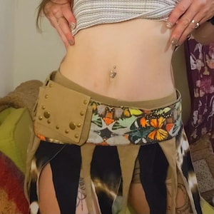 May include: A tan utility belt featuring a rectangular pouch with brass-colored studs and a wide band decorated with a colorful butterfly pattern. The belt includes long, dangling fabric strips in black and tan, hanging from the waistline.