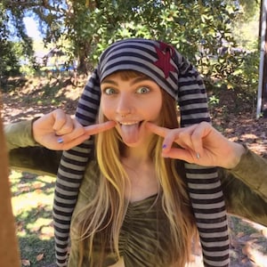 May include: A long, striped beanie hat featuring alternating dark gray and light gray horizontal bands, with a prominent dark red star-shaped patch attached to the side and two long, dangling ear-like extensions hanging down on both sides.