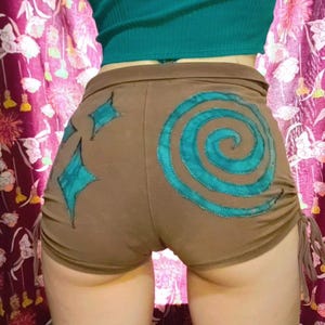 May include: Brown shorts with teal spiral and starburst designs. The shorts have drawstring sides and are paired with a teal crop top. The shorts are a soft, stretchy material.