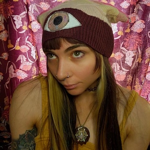 May include: A knit beanie with an eye design. The hat features a beige and burgundy color scheme, with a large eye patch sewn on the front. The eye has a white outline, a light brown iris, and a black pupil. The hat is a unique accessory.