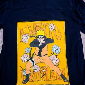 May include: Navy blue button-down shirt with a large yellow patch featuring a Naruto Uzumaki graphic. The graphic includes the character, flowers, and the words "NARUTO UZUMAKI". The shirt has long sleeves.