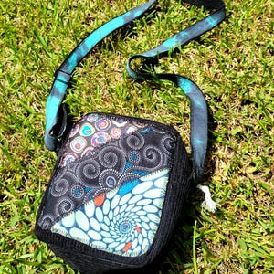 May include: A square crossbody bag featuring a patchwork front with black fabric, a spiral pattern in light blue and white, and a circular multi-colored design. The bag has a long, adjustable strap with a blue and black tie-dye pattern.