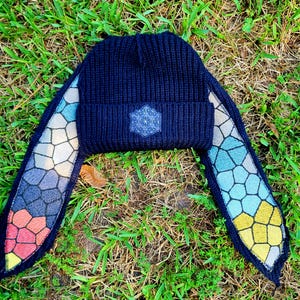 May include: A dark navy blue ribbed knit beanie featuring long, dangling side flaps. The front of the beanie displays a white geometric flower of life emblem. The side flaps have a multicolored stained glass pattern in red, blue, and yellow.