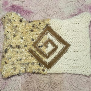 May include: A rectangular, two-toned pillow with a textured, beige and cream knit design. A brown, stitched square with a spiral pattern is centered on the pillow. The pillow is on a pink, textured surface.
